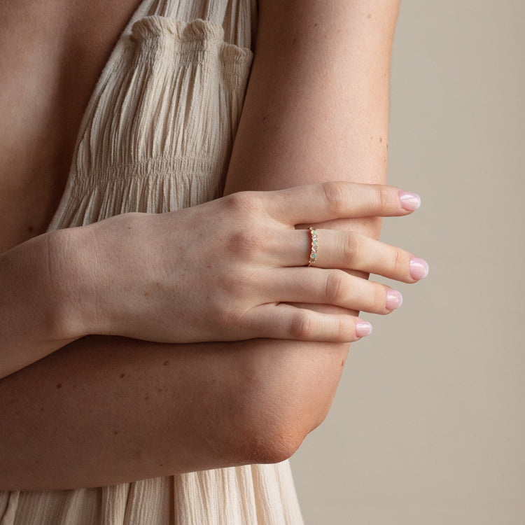 A person crosses their arms, displaying the 14k Australian Opal and Baguette White Sapphire Ring (sample size 7) on manicured fingers with light pink polish. They wear a sleeveless beige dress against a neutral background.