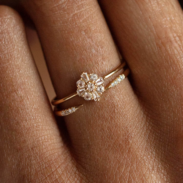 A close-up of a hand wearing two delicate gold rings: the 14k Open Front Stacking Ring with a cluster of small diamonds arranged like a flower, and a simple recycled gold band with tiny embedded diamonds.