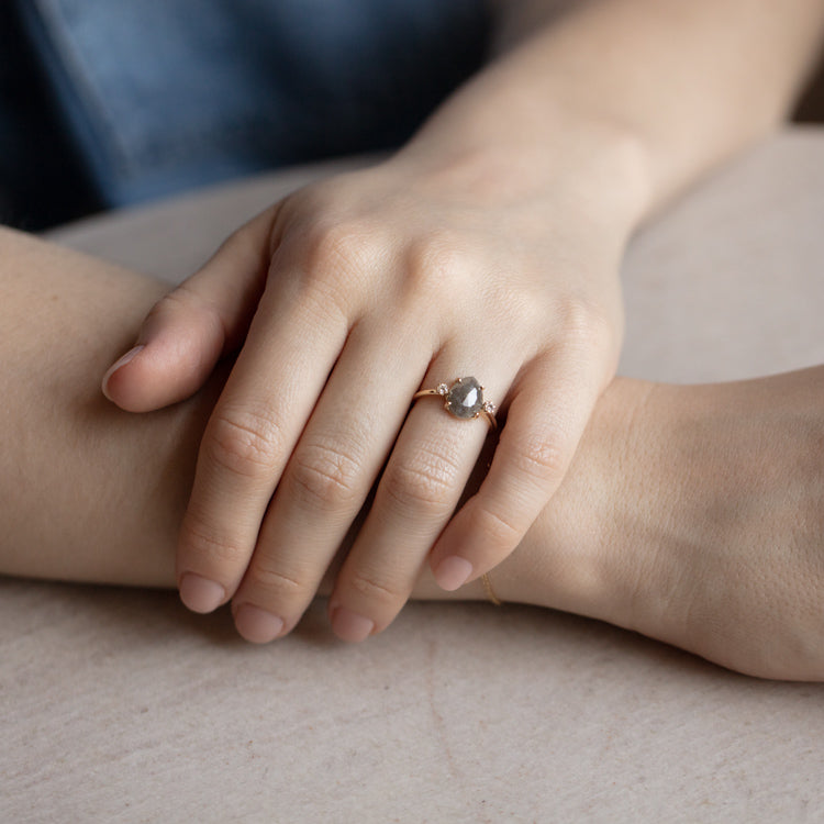 A close-up of a person’s hands resting on a surface, wearing the Eternal Grey Diamond Ring—a delicate yellow gold band with a pear-shaped grey diamond. Their nails are neatly manicured with a natural finish.