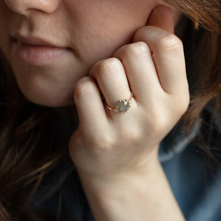 A close-up of a woman’s hand near her face, highlighting the Clair de Lune Grey Diamond Ring—a 14k gold band with a round rose cut diamond. Her lips, part of her chin, and long brown hair are also visible.
