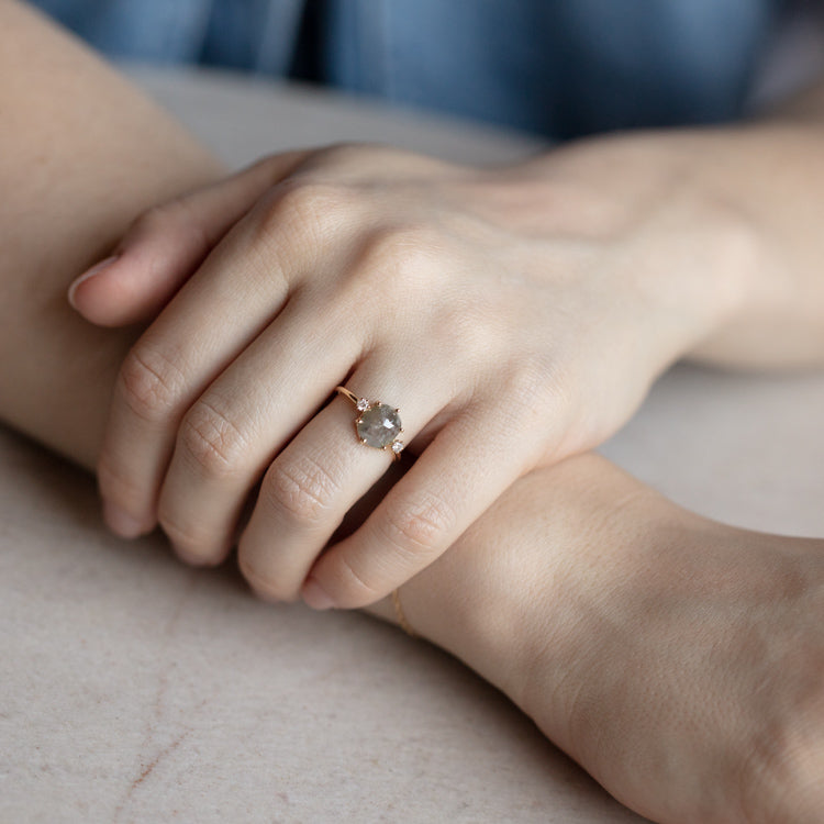 A close-up of two hands on a light surface, one adorned with the Clair de Lune Grey Diamond Ring—a delicate 14k gold piece featuring a round, rose-cut grey diamond.