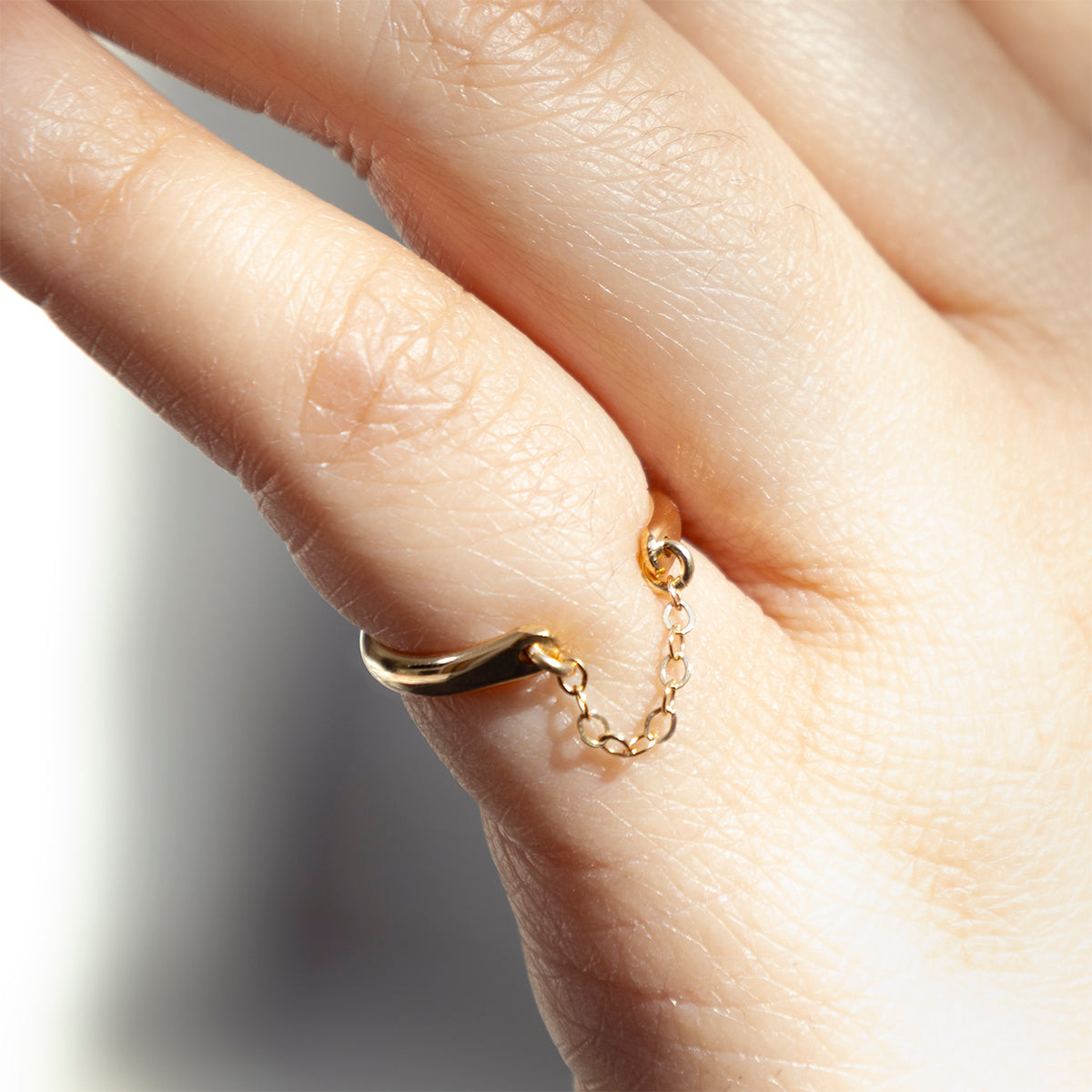 A close-up of a hand wearing the Glamour Ring, a unique 14k gold filled chain drop ring with a delicate chain wrapping around the finger, set against a softly lit background.