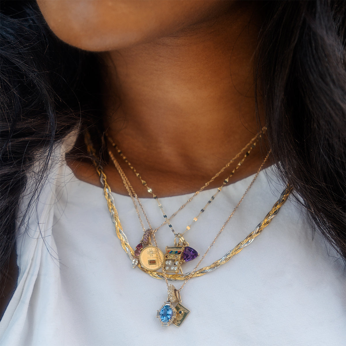 A woman in a white top wears layered gold necklaces, featuring Family Gold’s Solid Gold Trillion Birthstone Charm, a blue gem, locket, and purple stone pendants. Her dark wavy hair enhances the elegant style.