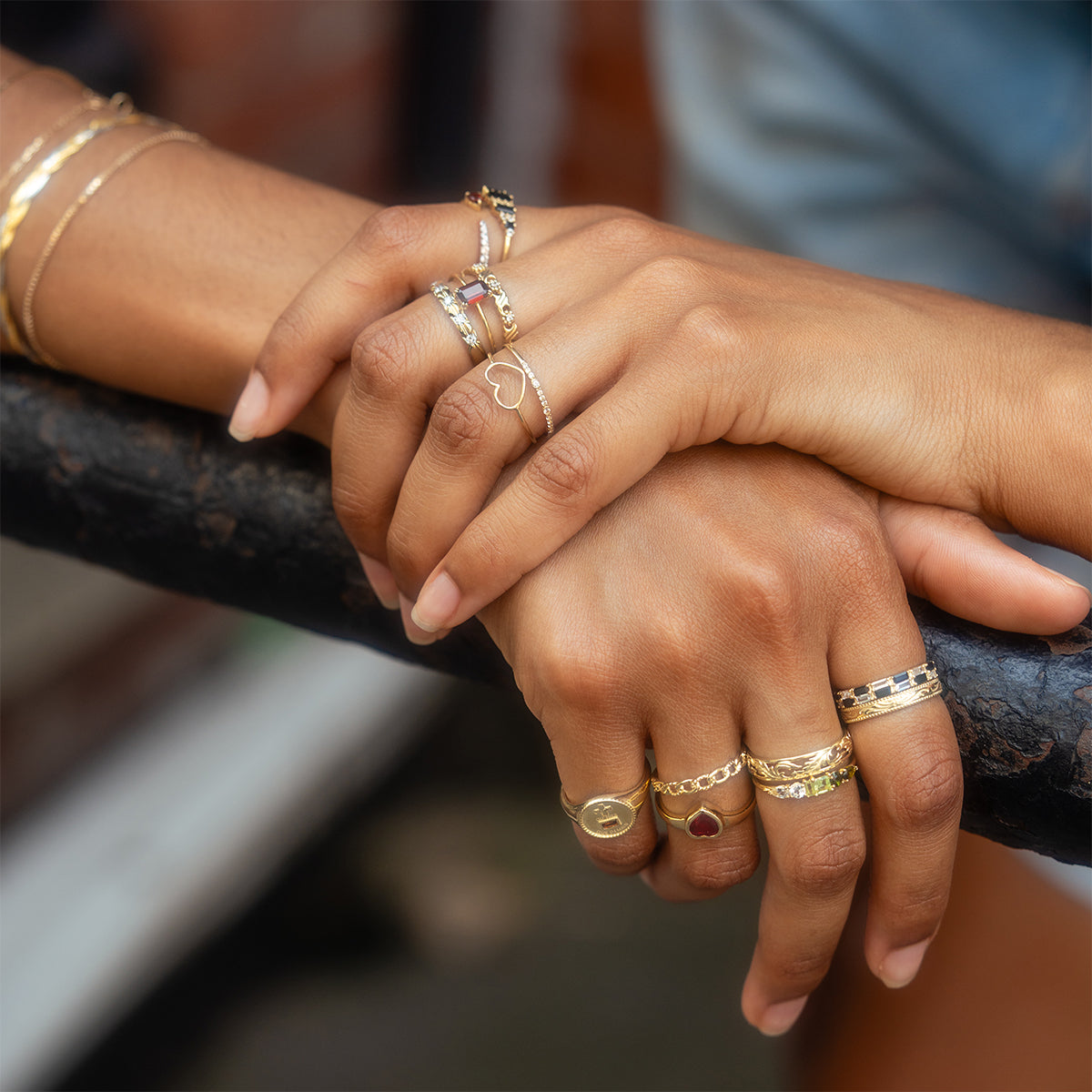 Two hands rest on a black metal railing, each adorned with gold jewelry. A Solid Gold Delicate White Sapphire Stacking Band adds subtle elegance, complementing a collection of rings and bracelets on naturally manicured nails.