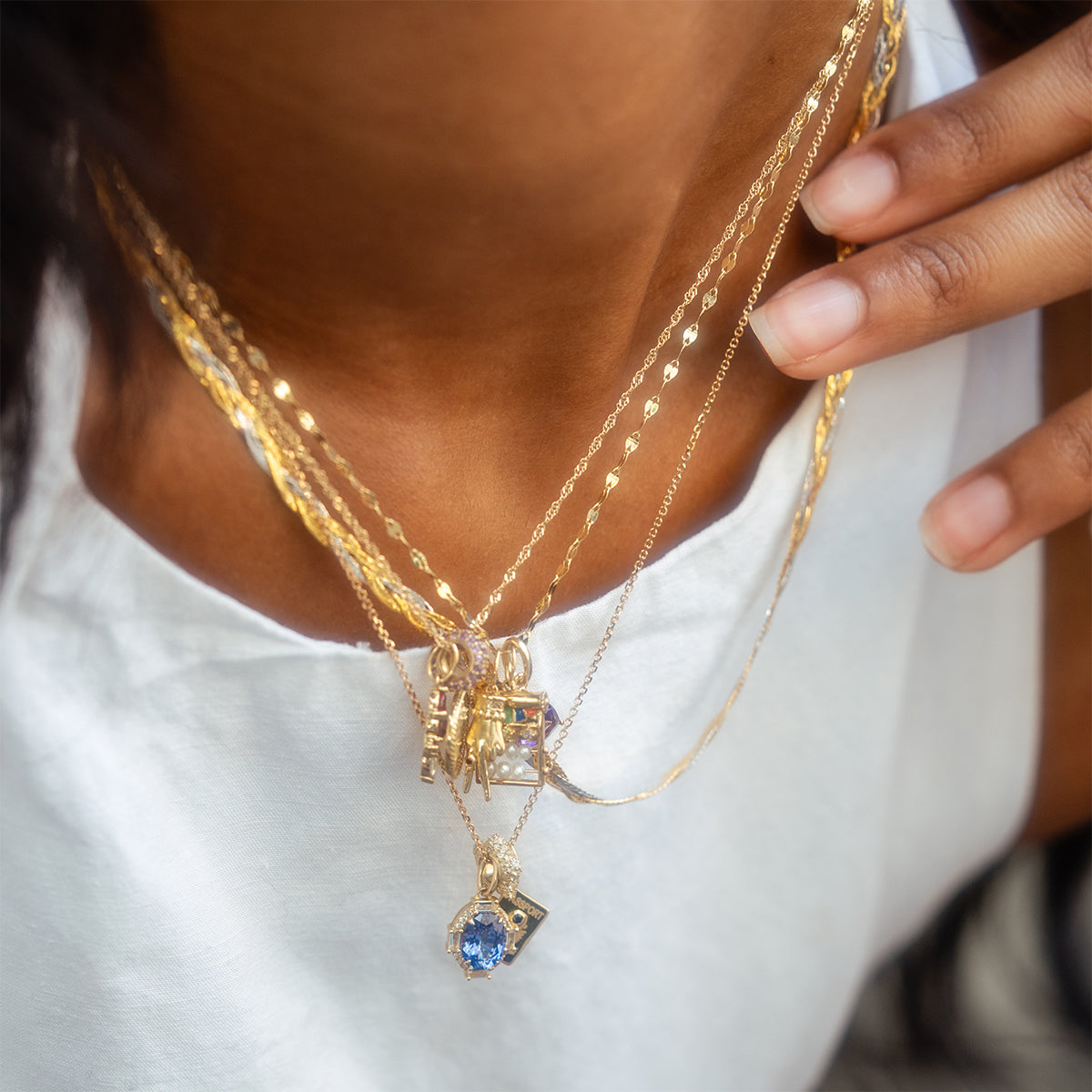 A close-up of a person wearing a white shirt and layered gold necklaces, featuring the Solid Gold Vintage Inspired Birthstone Charm with a blue gemstone, as their hand gently touches their neckline.