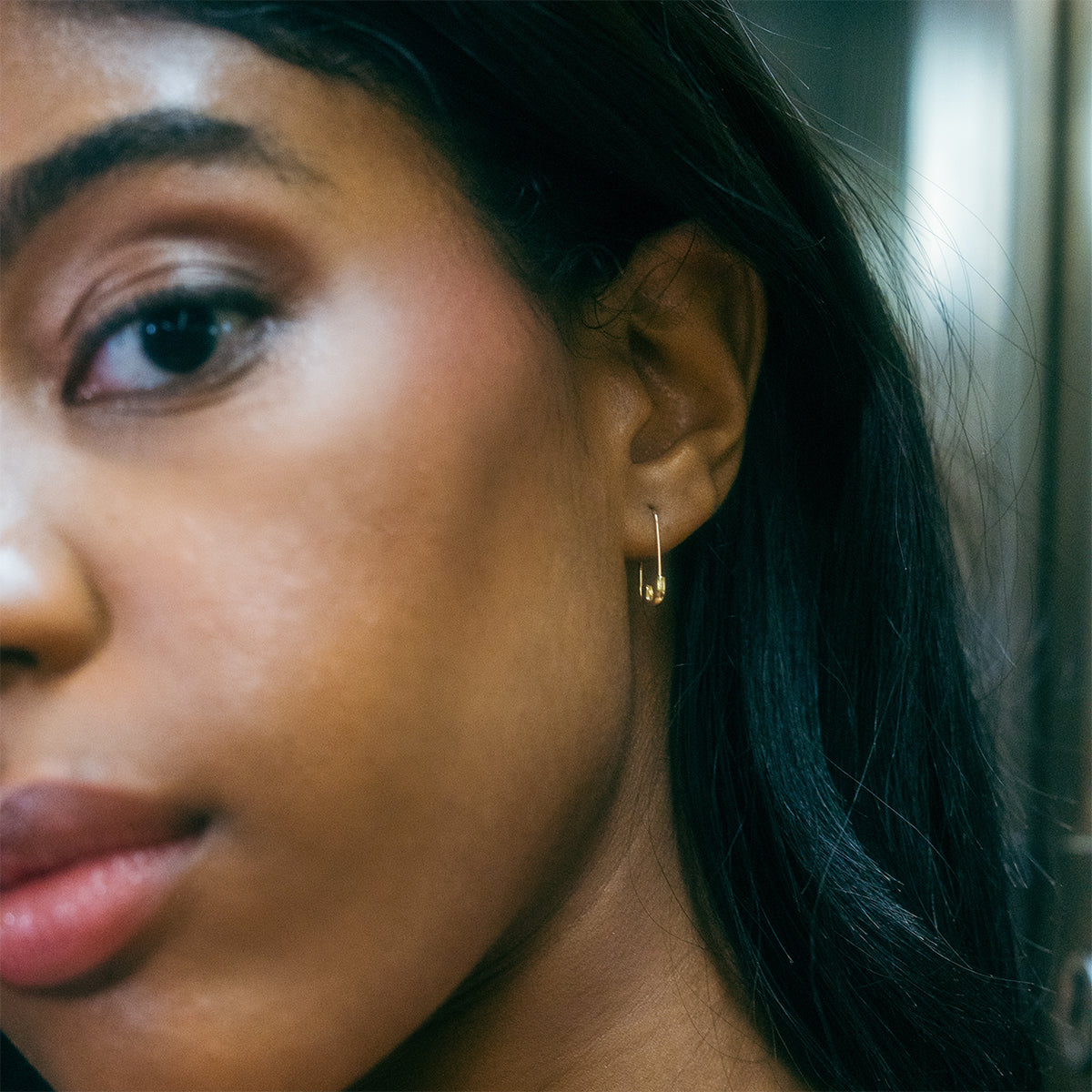 Close-up of a young woman’s face, focusing on her left ear adorned with a Solid Gold Safety Pin Earring. She has smooth skin, straight dark hair, and a calm expression against a softly blurred background.