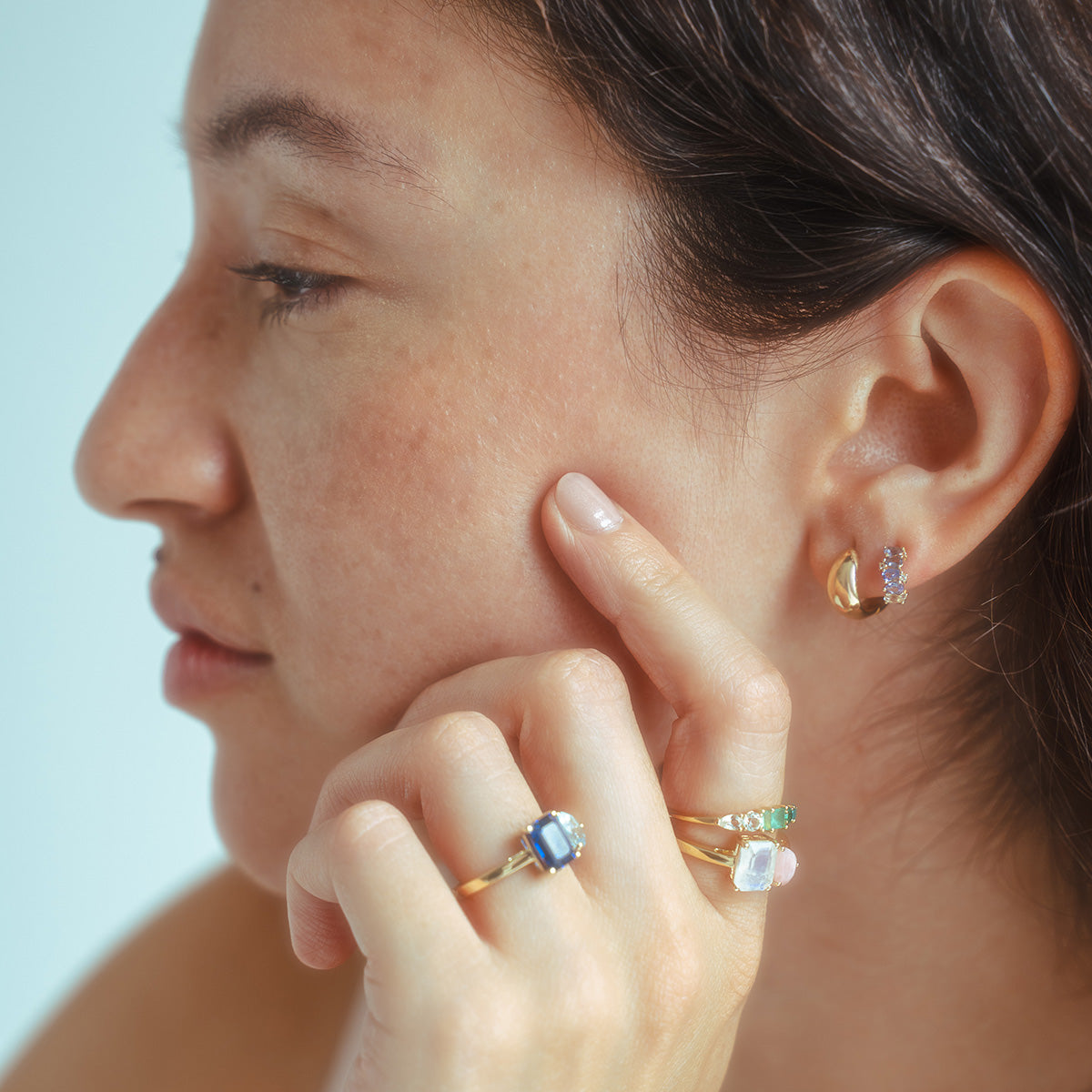A woman with medium skin tone and dark hair is shown in profile, wearing multiple gold rings with colorful gemstones and Solid Gold Chunky Huggie Hoops on her ears. She touches her face gently, appearing thoughtful.