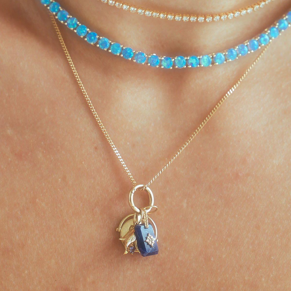 A close-up of a person’s neck wearing three necklaces: a thin gold chain with blue and gold charms, a choker with round blue stones, and a delicate Solid Gold Birth Flower Bud Charm pendant.