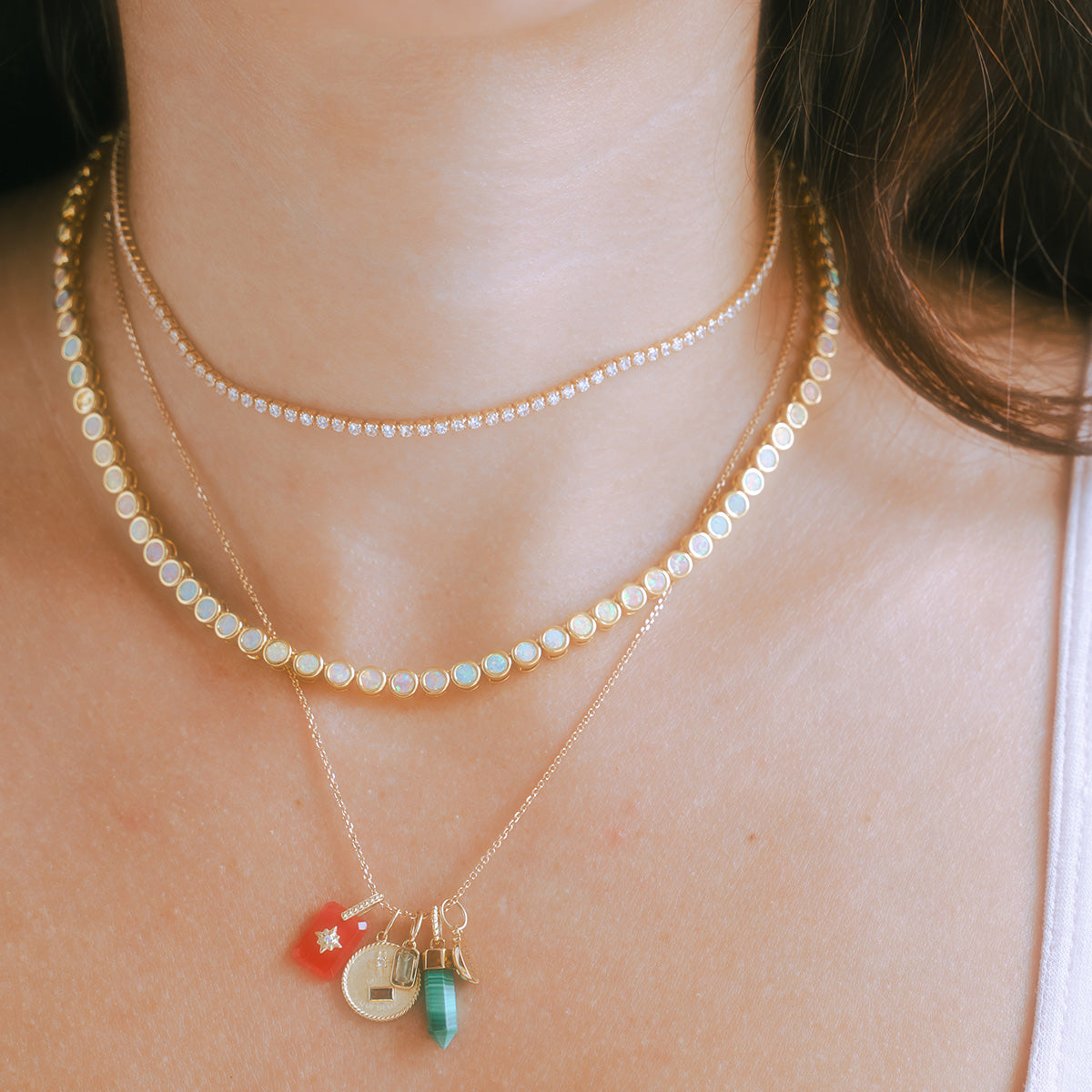 A close-up of a womans neck wearing four layered gold necklaces, featuring the Solid Gold Crystal Amulet Charm for Manifestation with natural crystals, small diamonds, opal-like stones, and pendants such as a red charm and green stone.