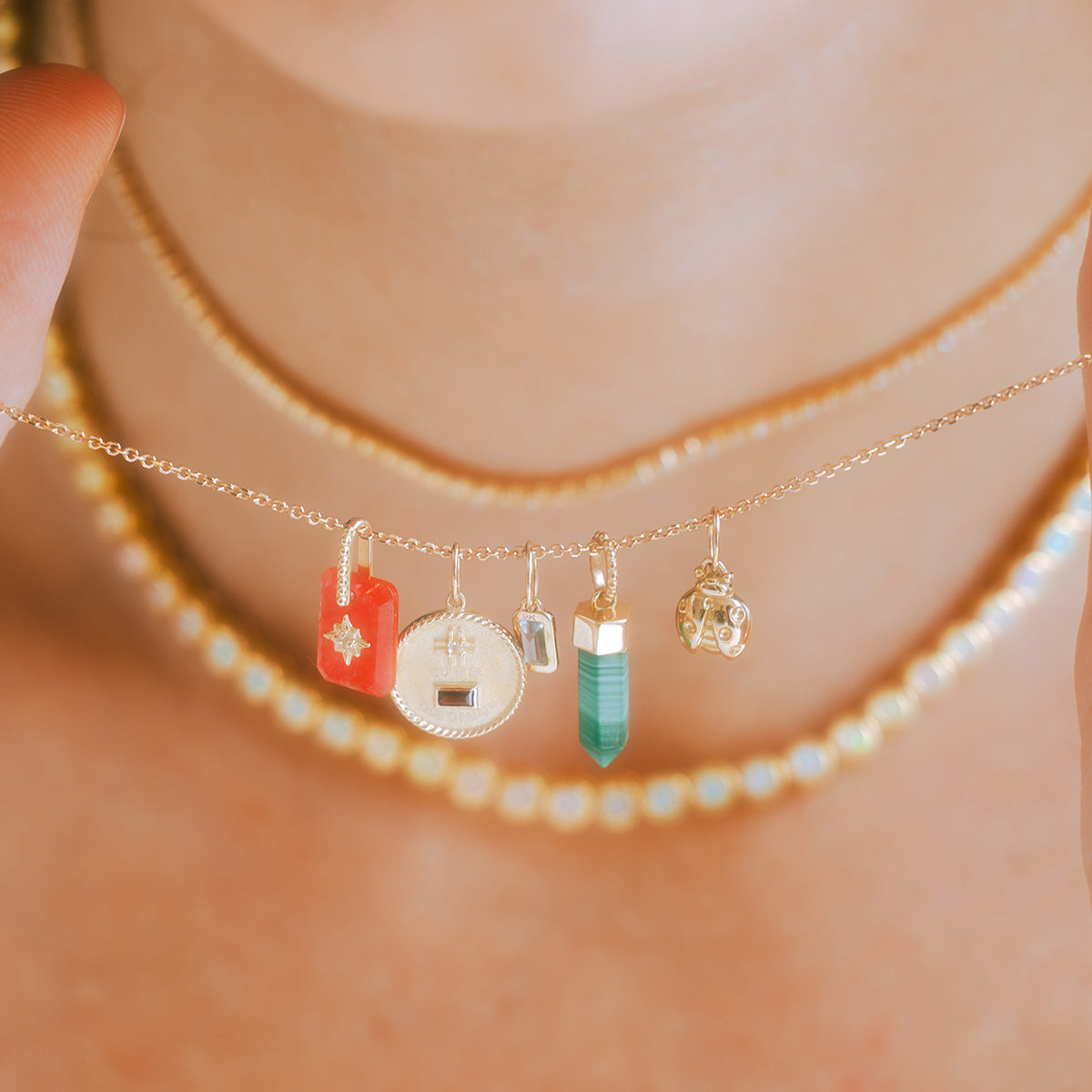 A close-up shows a person holding the Solid Gold Crystal Charm for Manifestation, featuring five colorful charms—a red tag, coin, crystal, green stone, and gold crystal—while wearing two natural crystal beaded chokers.