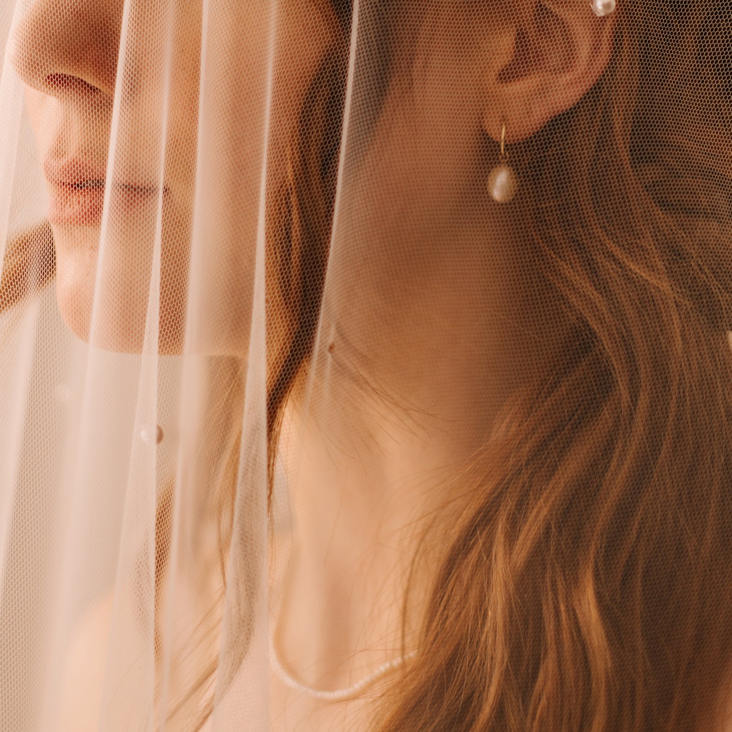 A close-up of a woman with wavy hair wearing Solid Gold Elegant Pearl Charm Huggies and a sheer white veil draped over her face and shoulder, creating a soft, delicate atmosphere.