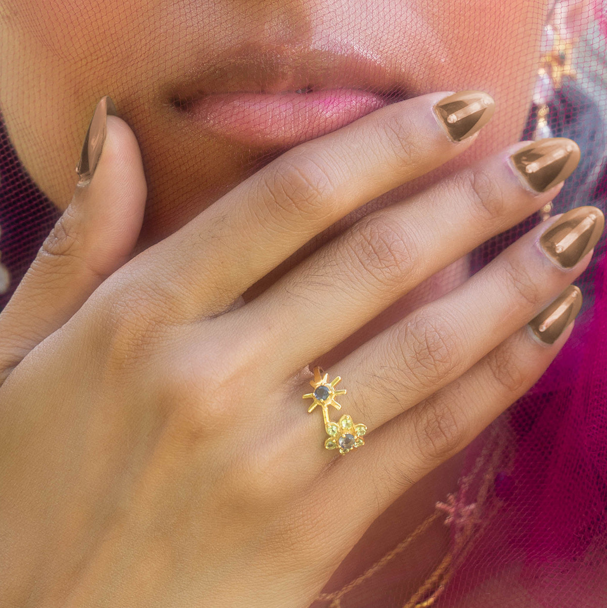 A close-up of a hand with metallic nude nail polish touches the face, highlighting the Sungoddess Ring Sample Size 6—an 18k yellow gold plated ring with a sun-shaped design and Iolite gemstones. Lips and part of the nose are visible in the background.