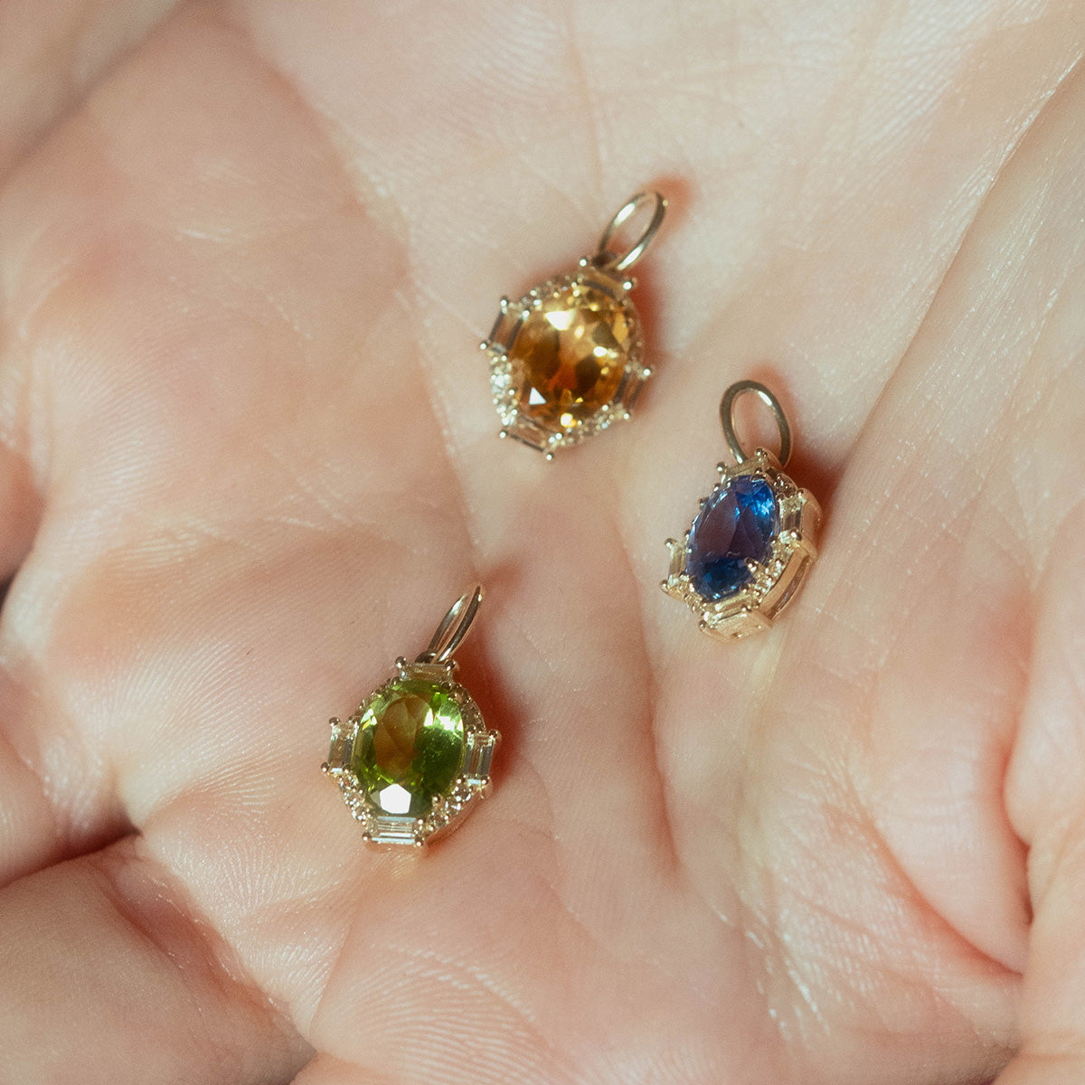 A hand displays three Solid Gold Vintage Inspired Birthstone Charms featuring faceted oval gemstones—yellow, green, and blue—each surrounded by sparkling clear stones in an ornate setting.