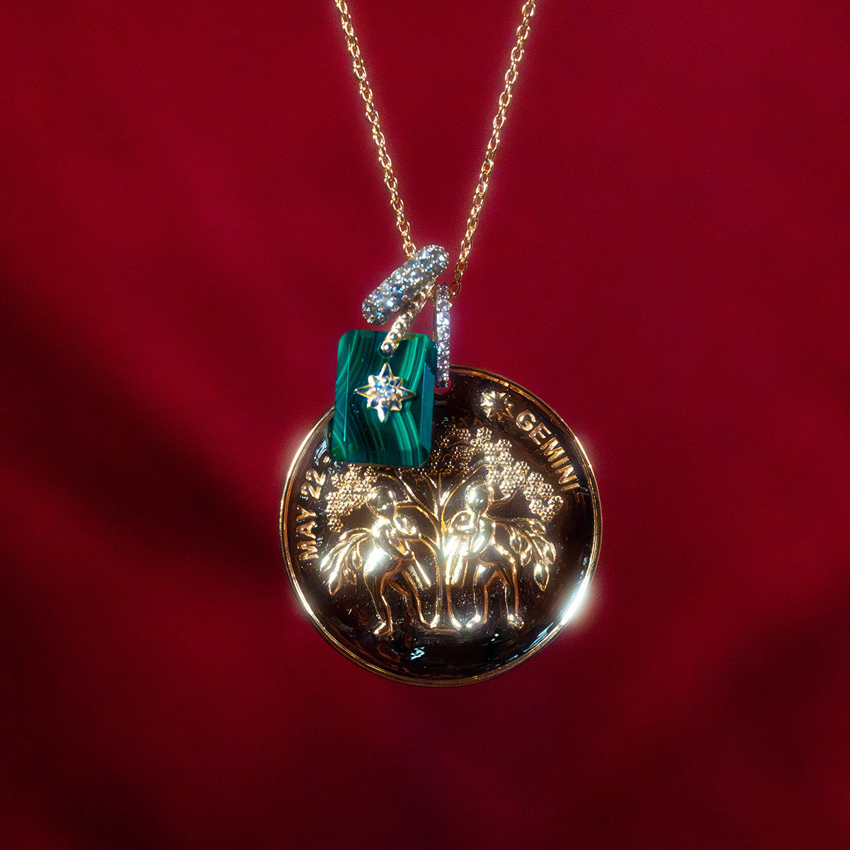 The Vintage Inspired Zodiac Coin Necklace features a gold vermeil chain with two Gemini figures, a green rectangular charm, and a diamond-encrusted G, set against a deep red backdrop.
