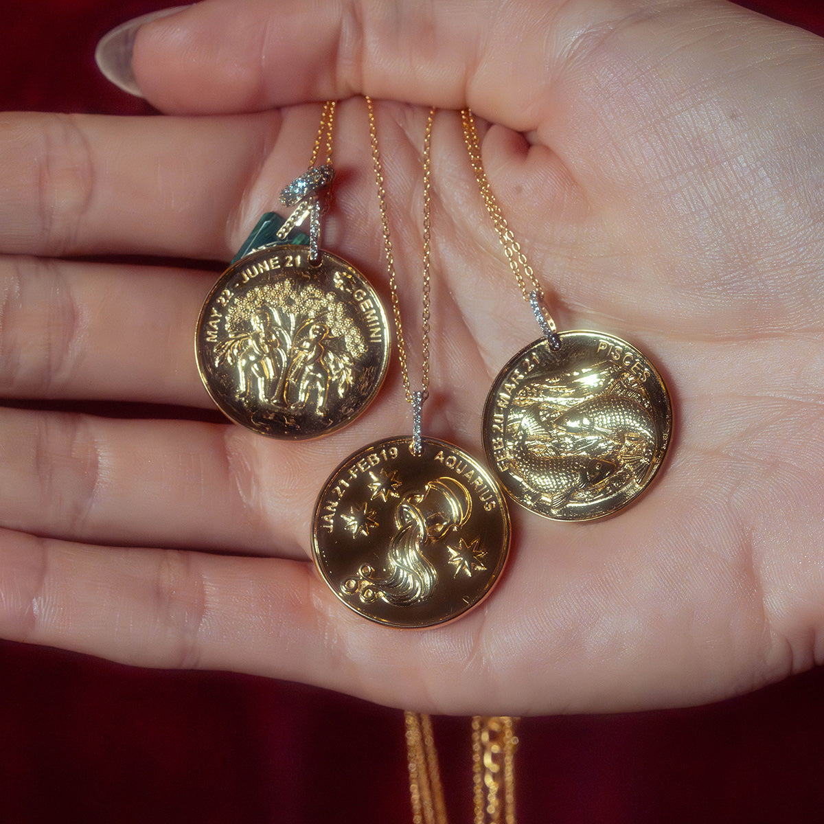 A hand holds three Vintage Inspired Zodiac Coin Necklaces, each with intricate gold designs engraved with the names and dates of Gemini, Aquarius, and Capricorn, adding a touch of vintage charm.