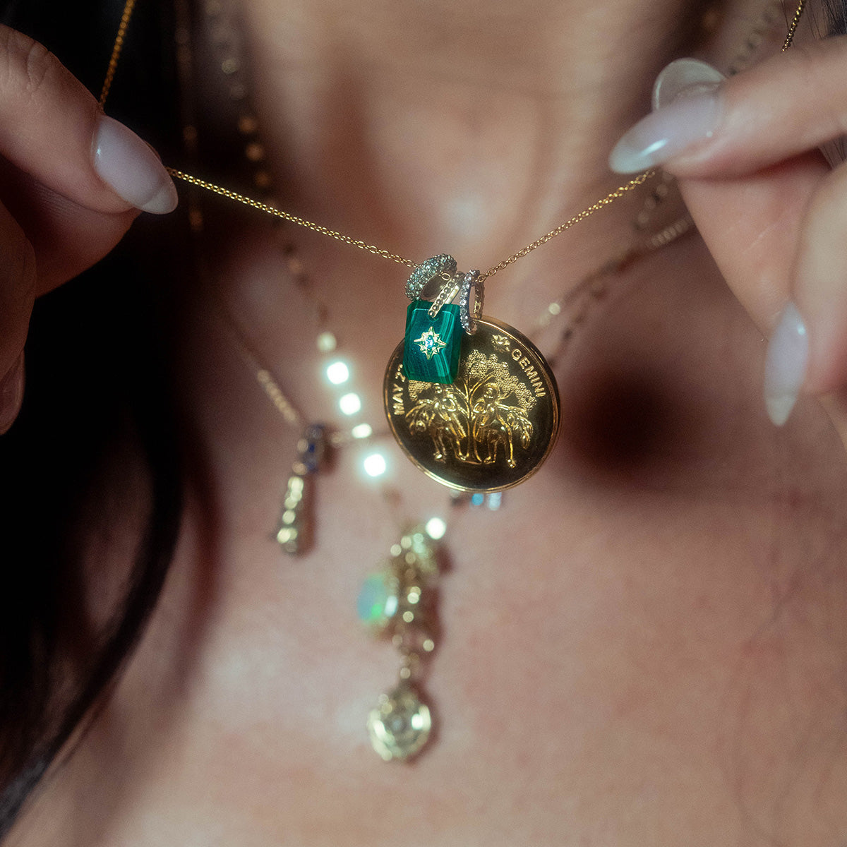 A person holds up the Vintage Inspired Zodiac Coin Necklace, featuring a gold vermeil round pendant engraved with palm trees and GERMANY, accented by a green crystal charm and rhinestone. More layered vintage charm necklaces appear in the background.