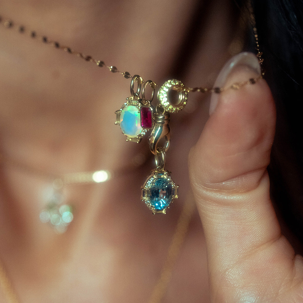 A hand holds the Solid Gold Vintage Inspired Birthstone Charm with three gemstone charms—blue, red, and iridescent—while another vintage-inspired pendant shimmers softly in the blurred background.