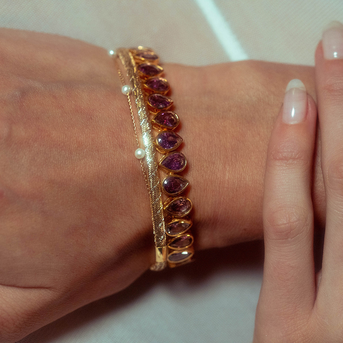 A close-up of a hand wearing two bracelets—a Solid Gold Mixed Metal Mesh Bracelet with purple gemstones and another with small pearls—against a light background, while another hand gently touches the wrist.