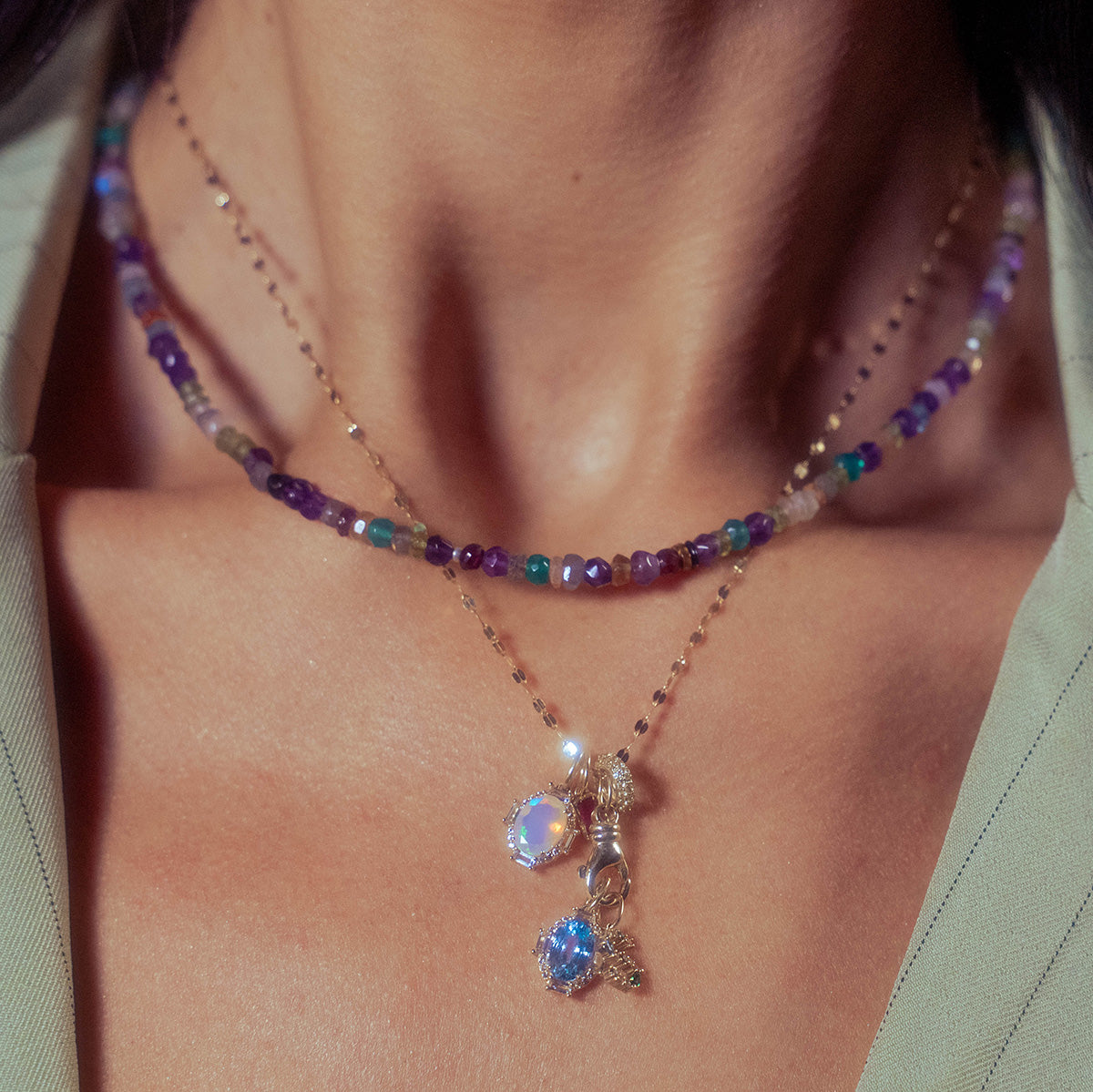 A close-up of a womans neck showcases two layered necklaces: one with colorful beads, and the other featuring the Solid Gold Vintage Inspired Birthstone Charm. She wears a light green top.