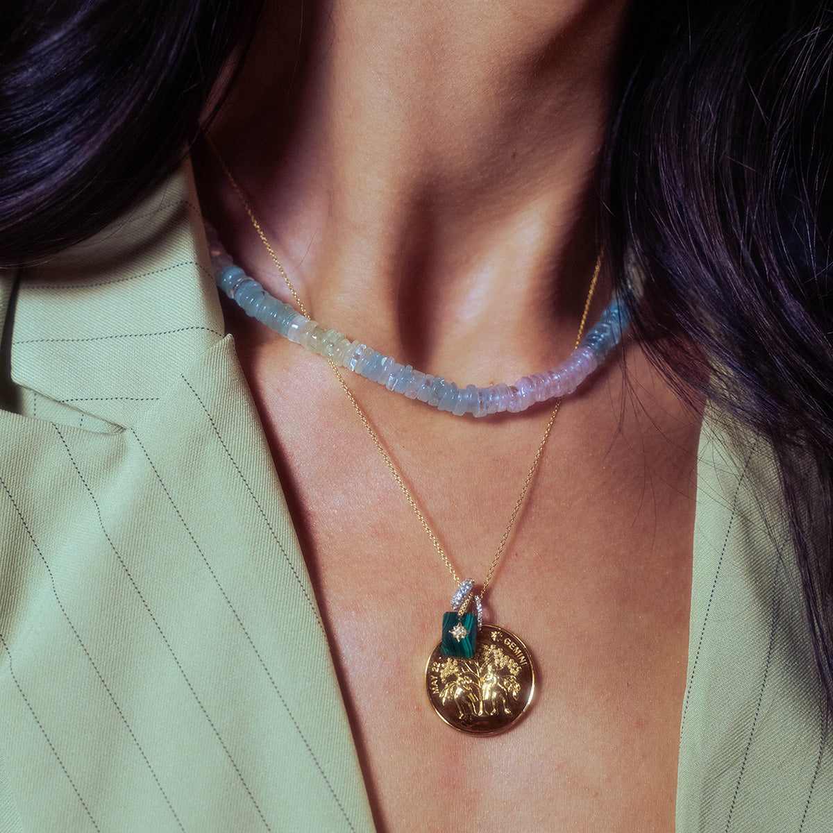 A close-up of a person wearing a light green pinstripe blazer, pastel beaded necklace, and two gold pendant necklaces—one featuring green and gold details, and the other is a Solid Gold Crystal Amulet Charm for Manifestation.