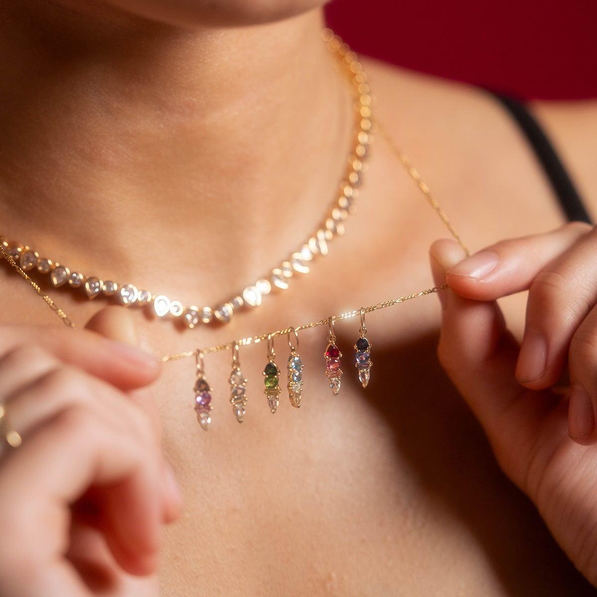 A person holds a gold necklace with six gemstone pendants, including a Solid Gold Modern Ombre Birthstone Charm, while wearing another gold necklace with small round accents. The blurred background highlights the stunning jewelry.