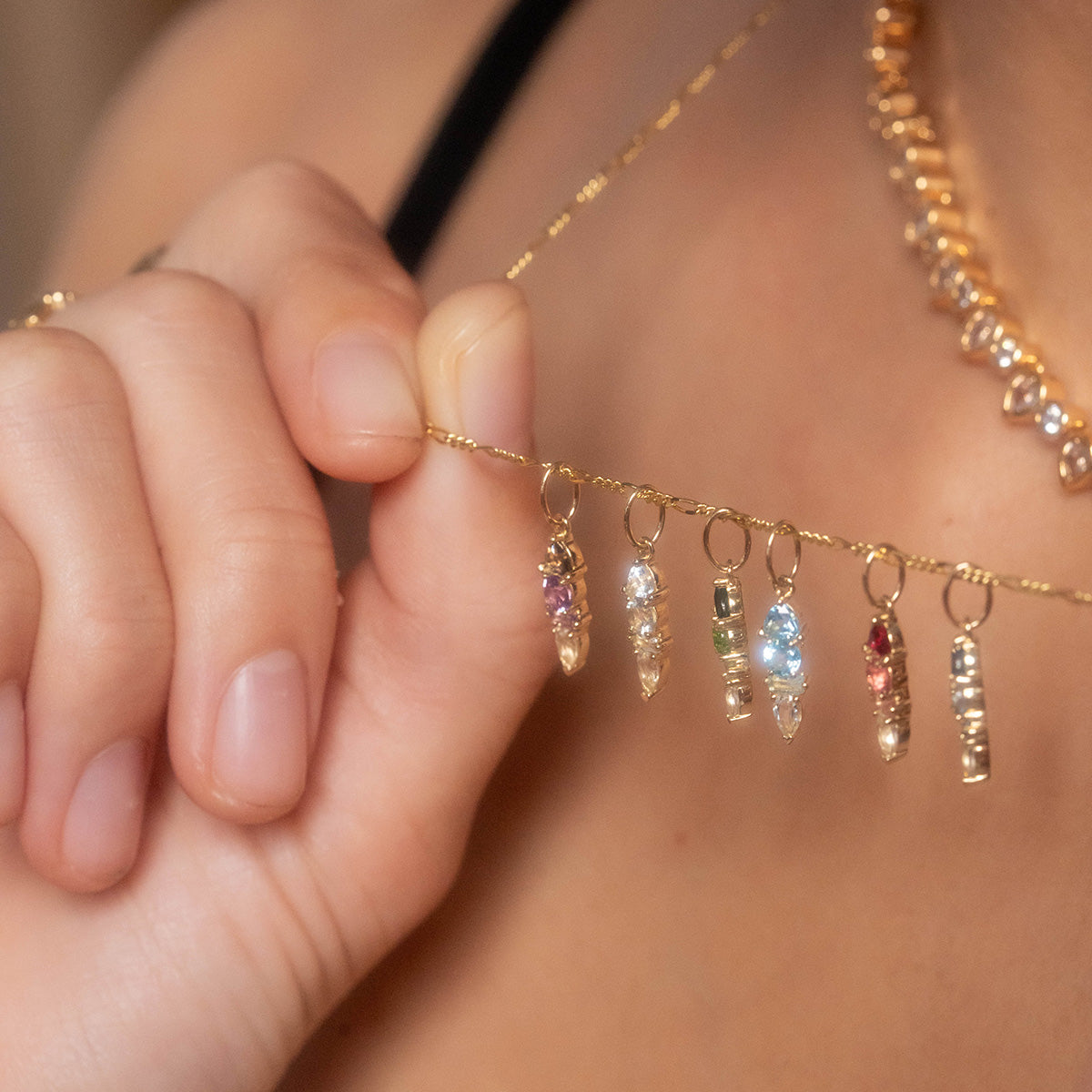 A close-up of a hand holding the Solid Gold Modern Ombre Birthstone Charm necklace, featuring multiple crystal pendants with various colored stones, including a birthstone charm, set against a beautifully blurred background.