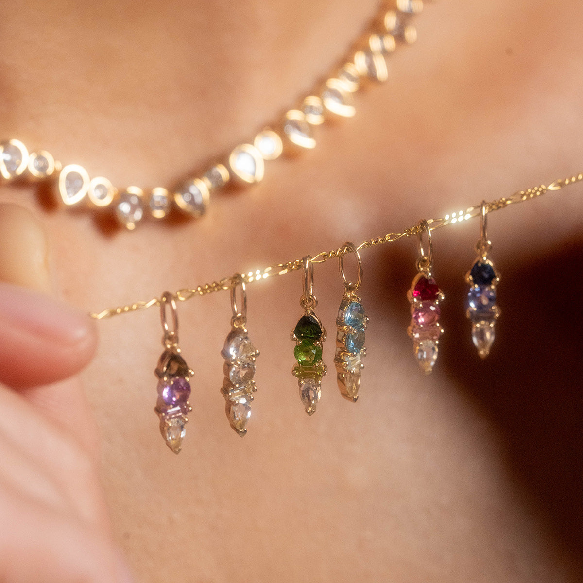 A person holds the Solid Gold Modern Ombre Birthstone Charm, featuring six colorful crystal pendants resembling birthstones. They wear a solid gold necklace with round and heart-shaped charms against a softly blurred background.