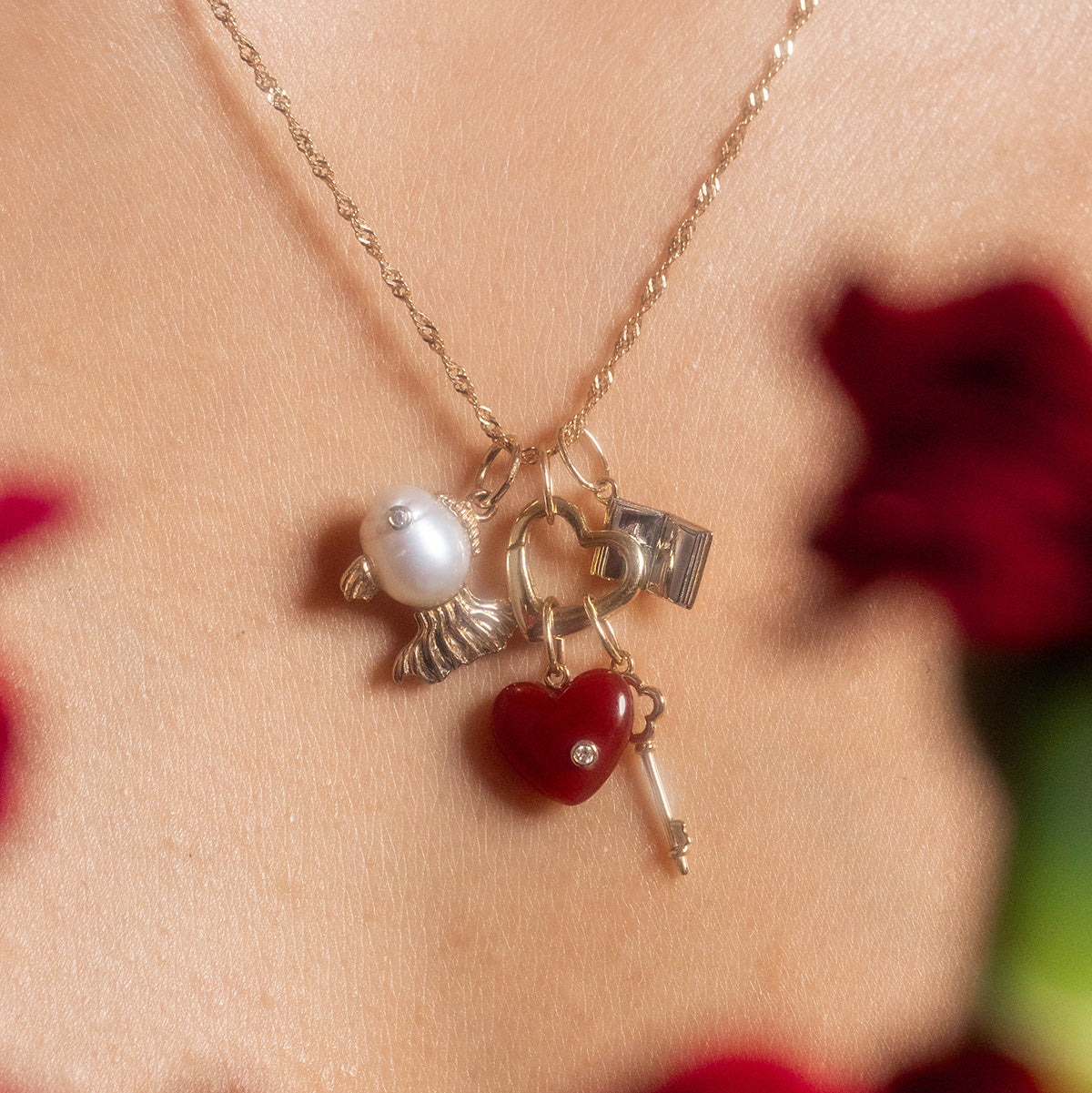 The Solid Gold Statement Charm necklace, crafted in 9k yellow gold, features four charms—a white bird, padlock, red heart with a gem, and small key—worn on the neck amid blurred red flowers.