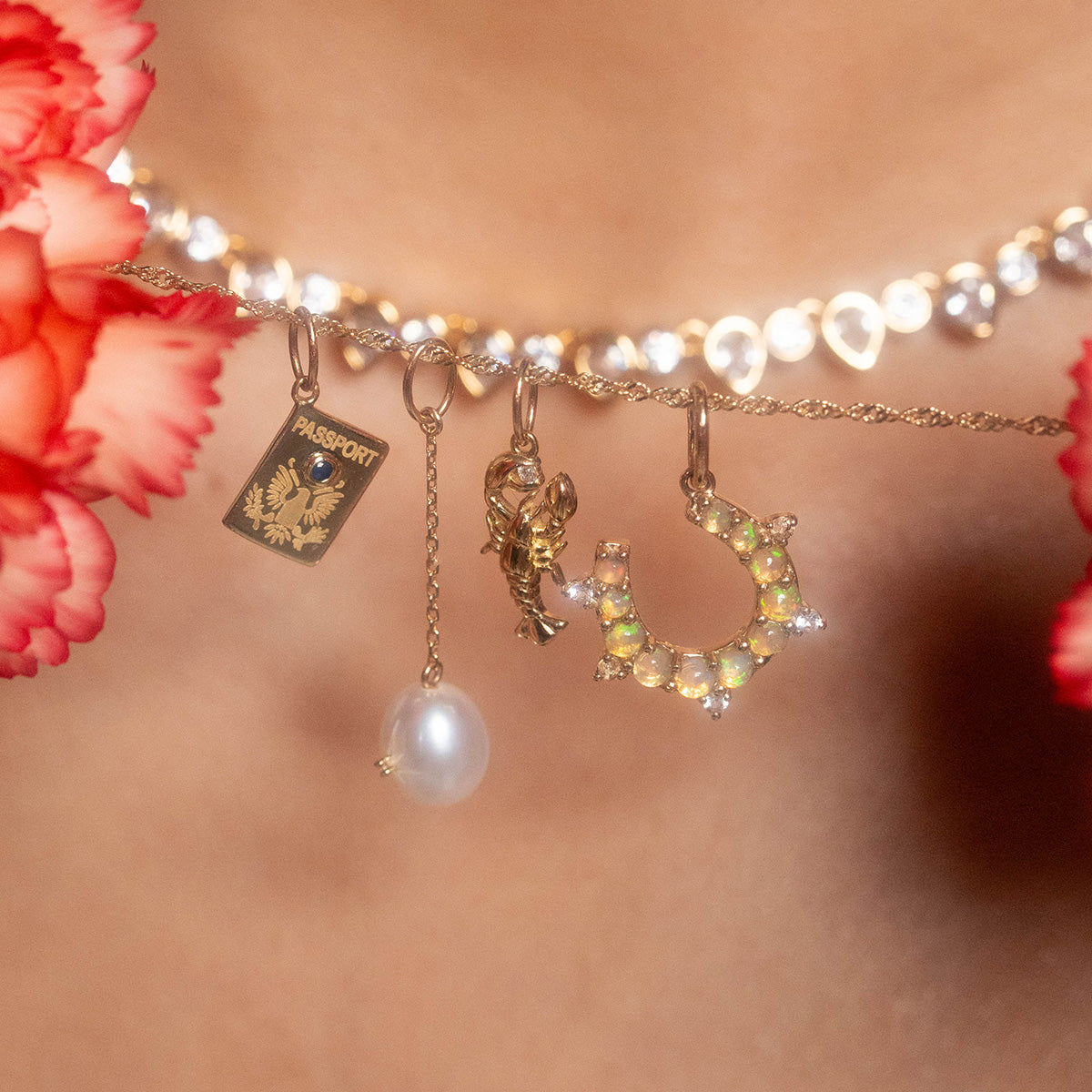 Close-up of the Solid Gold Celebration Charm necklace, featuring four engraved gold charms—a miniature passport, a pearl, a tiny lobster, and an opal-encrusted crescent—with blurred red carnations in the foreground.