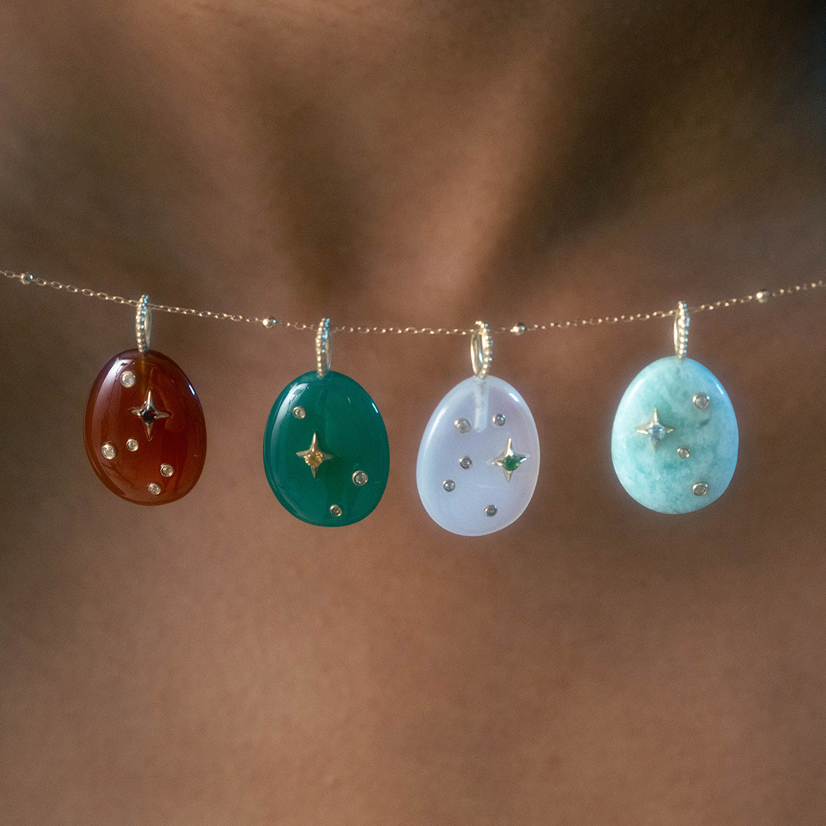 A close-up of the Solid Gold Zodiac Constellation Crystal Charm necklace, featuring four oval gemstone pendants—red, green, light purple, and light blue—each with gold star and dot designs on a delicate gold chain.