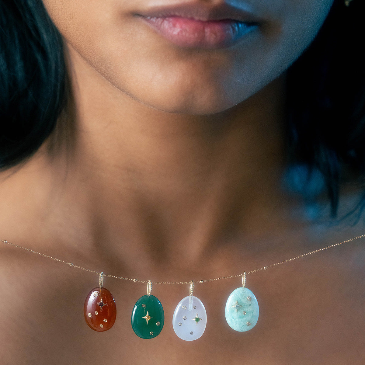 A close-up of a person’s neck showcases the Solid Gold Zodiac Constellation Crystal Charm, featuring four oval gemstone pendants and gold star accents on a delicate chain. The wearer’s lips are partially visible.