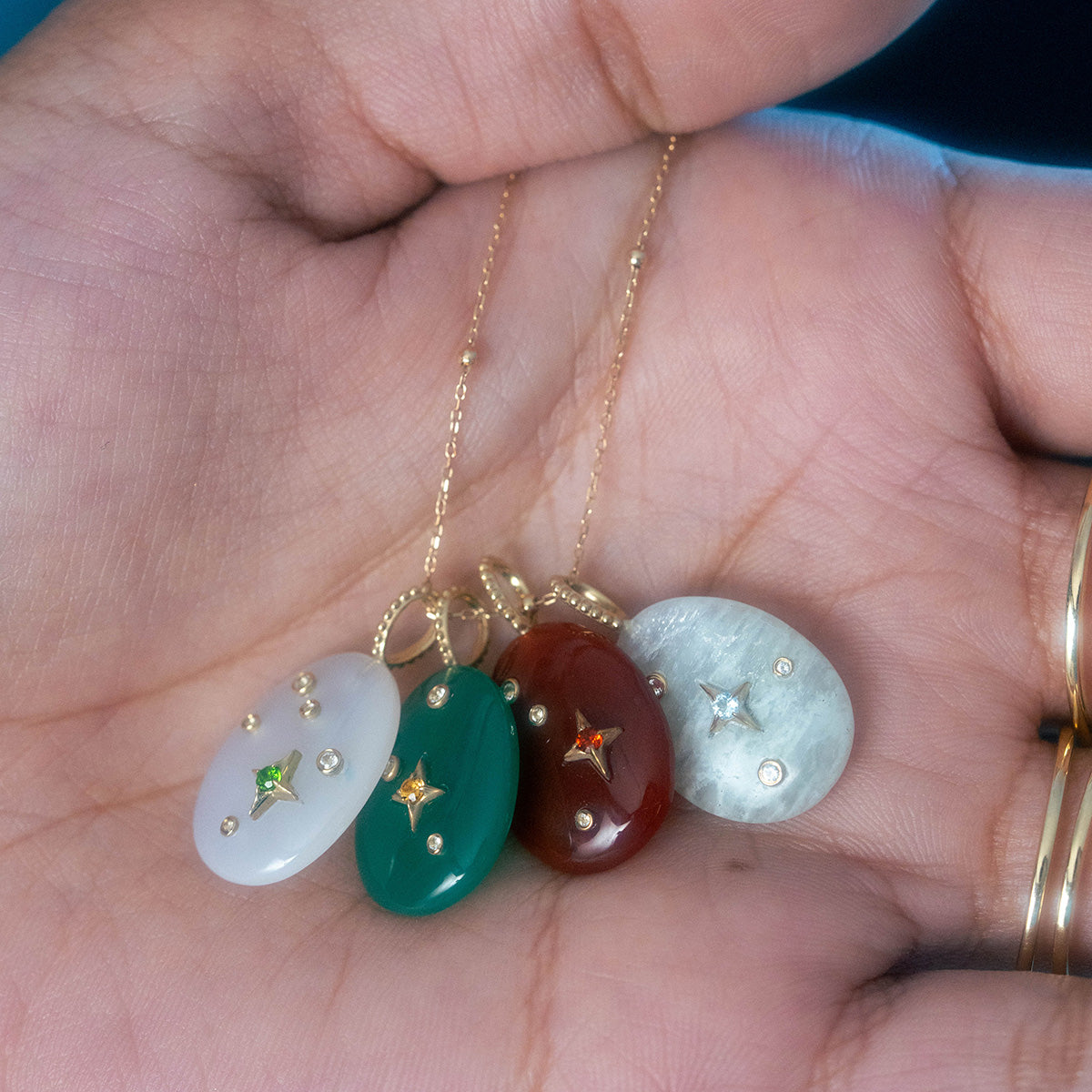 A hand holds four Solid Gold Zodiac Constellation Crystal Charms—oval pendants in white, green, red, and gray—each with a star-shaped gold setting and center gemstone, perfect as personalized zodiac or crystal jewelry.