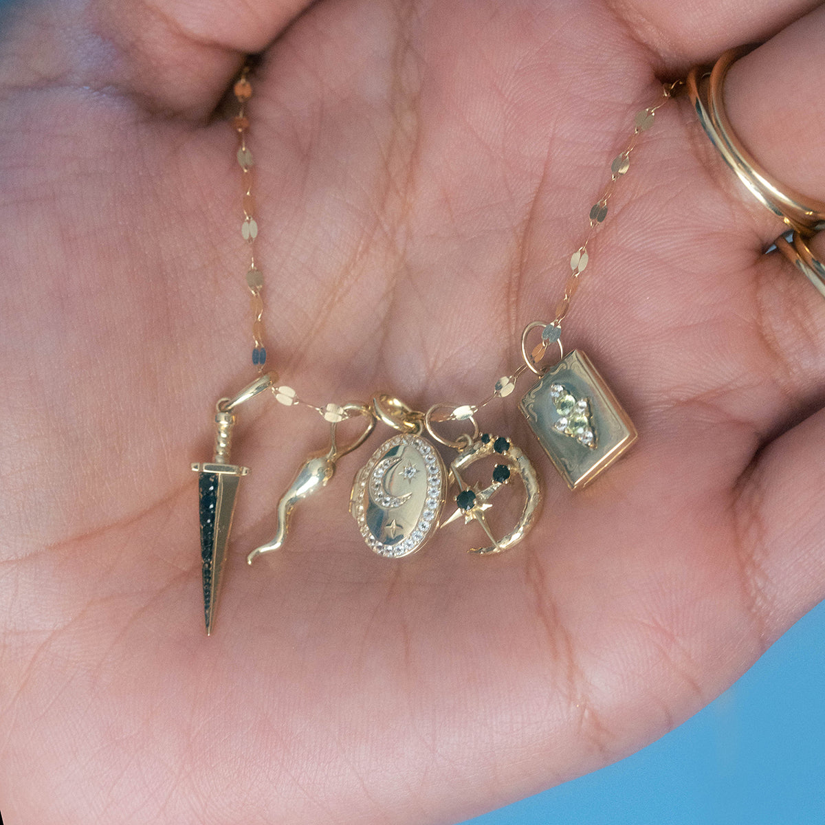 A hand holds a gold chain necklace featuring five unique charms, including the Solid Gold Amulet Charm. The wearer also has on two gold rings. This is a Local Eclectic exclusive.