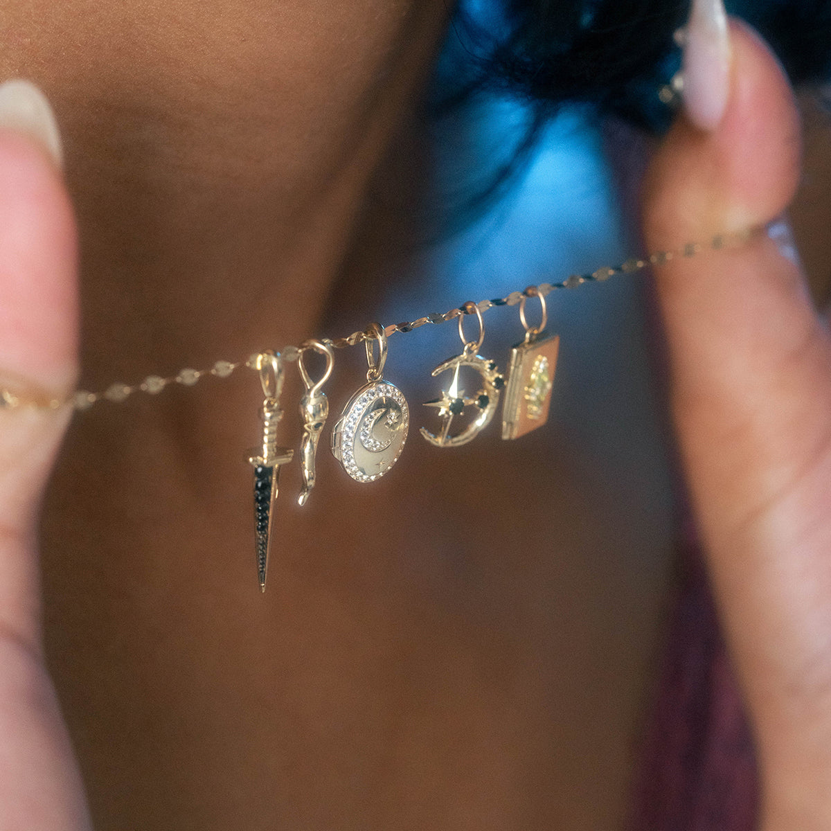 A person holds the Solid Gold Amulet Charm, a necklace featuring five pendants—dagger, clock, crescent moon with stars, planet, and book—exclusive to Local Eclectic. A softly blurred background highlights this unique jewelry piece.
