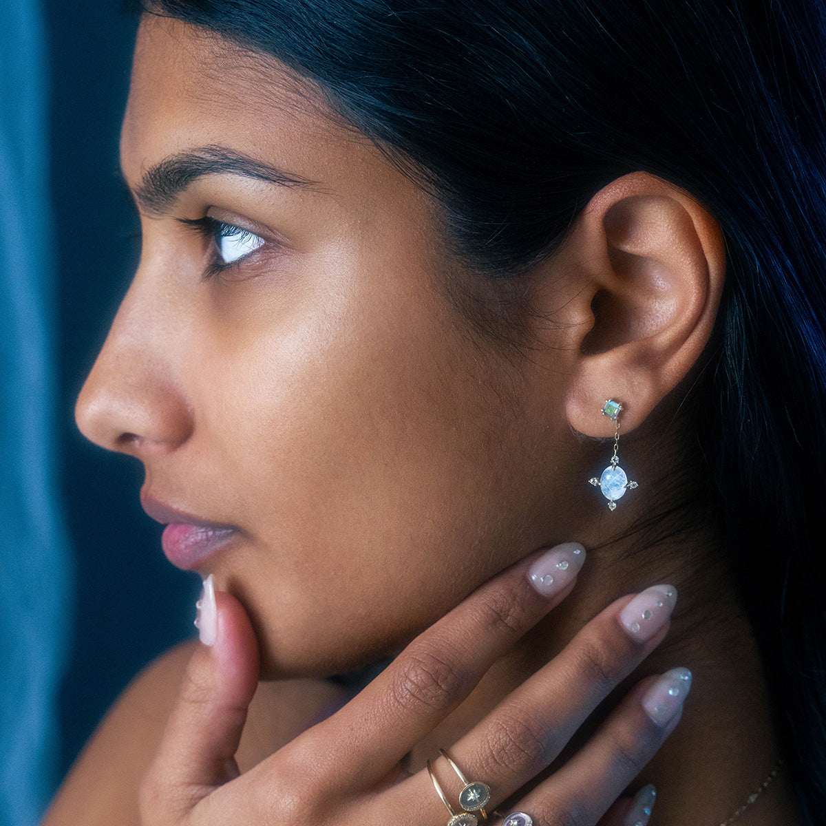 A person with smooth brown skin is shown in profile wearing Solid Gold Crystal Ball Moonstone Drop Earrings. Their hand, adorned with rings, gently touches their chin, displaying long, manicured nails.