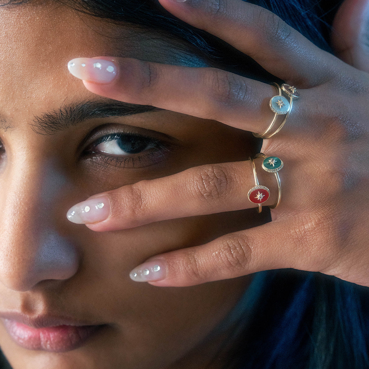 A person with pearl-adorned nails poses with their hand partially covering their face, showcasing three gold rings with colorful enamel star designs and a Solid Gold Crystal Crescent Moon Ring for Manifestation for a mystical touch.