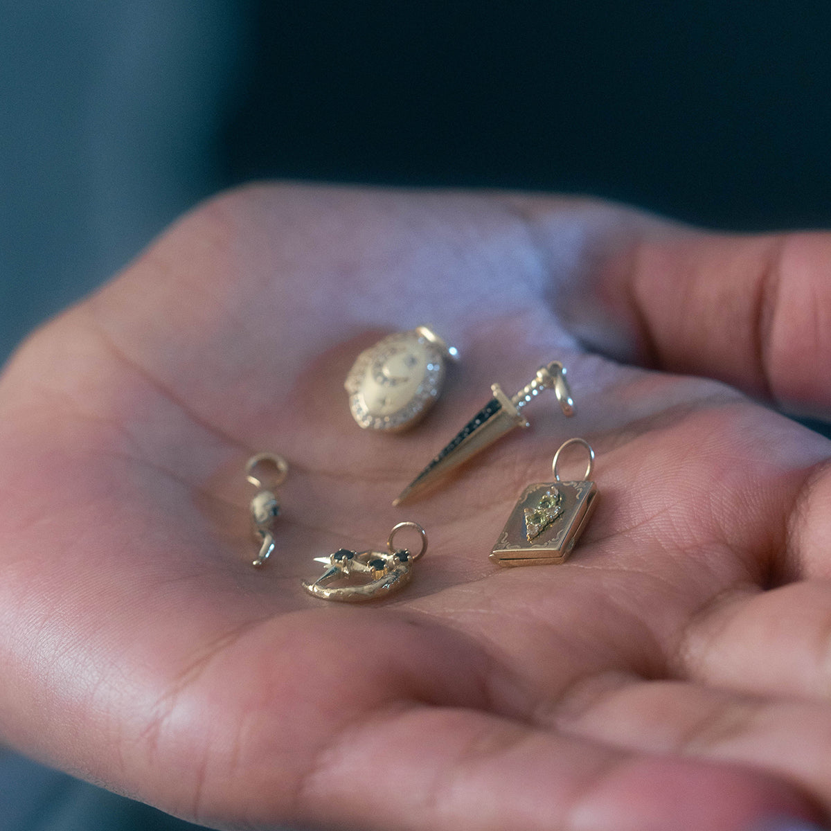 A hand holds five Solid Gold Amulet Charms—shield, sword, pistol, mask, and book—each intricately detailed, creating a truly unique local eclectic exclusive.