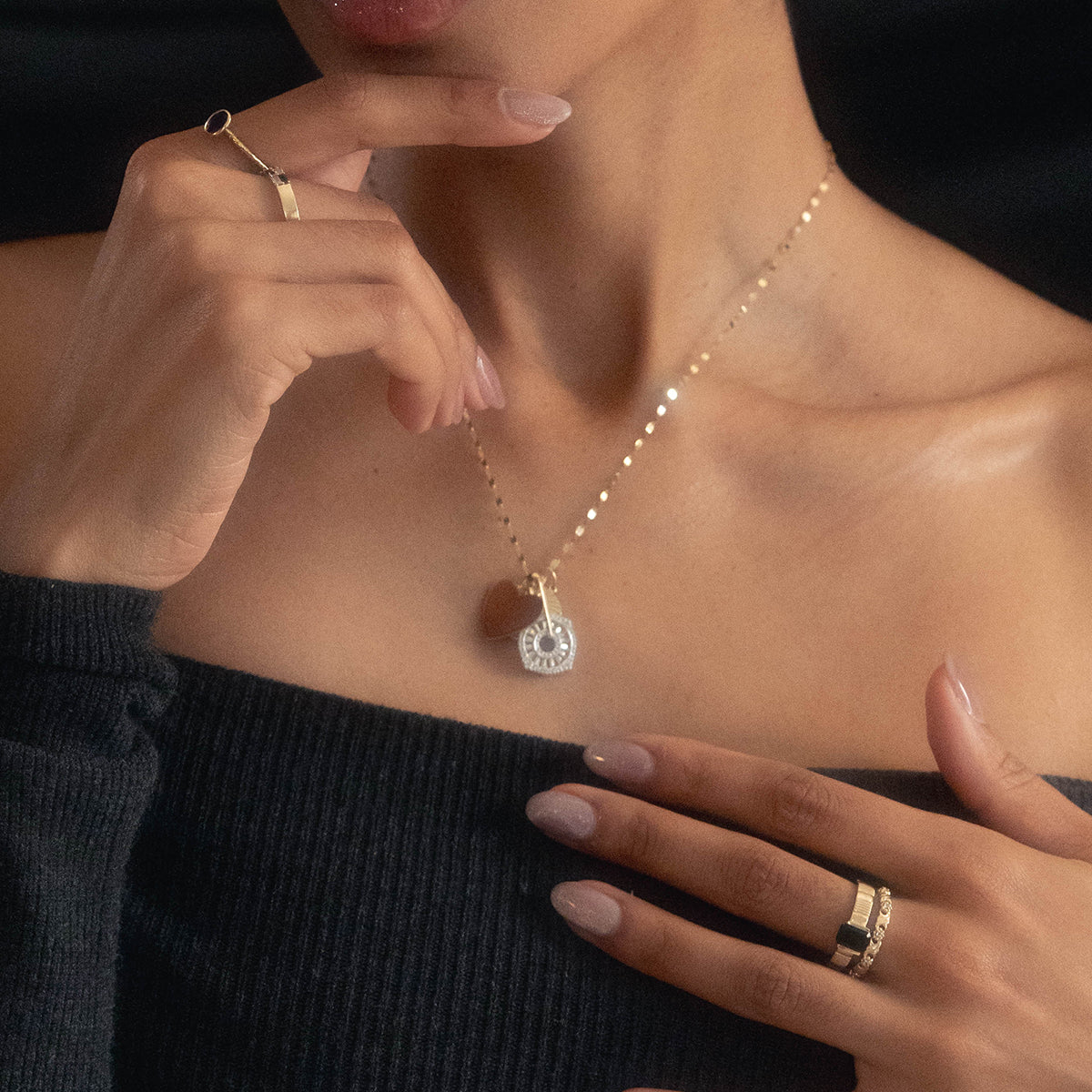 A woman wearing a solid gold vintage inspired sapphire floral band touches her lips and collarbone with manicured fingers, dressed in an off-the-shoulder black top against a dark background.