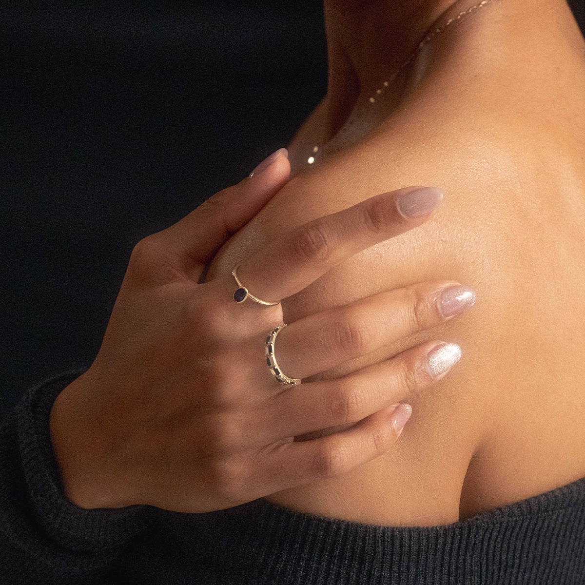 A person with manicured nails gently rests their hand on their bare shoulder, wearing the Solid Gold Amethyst Oval Ring from Local Eclectic and a delicate gold necklace, set against a dark background.