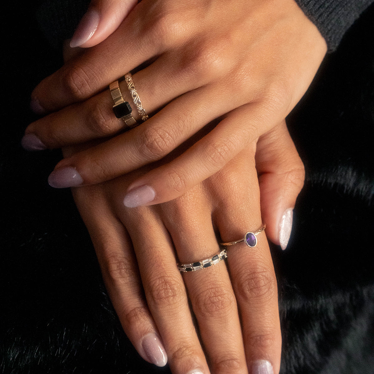 Two hands with manicured, light pink nails rest together, wearing three rings—a Solid Gold Black and White Checkerboard Band, a black agate ring, and one set with a small oval purple stone.