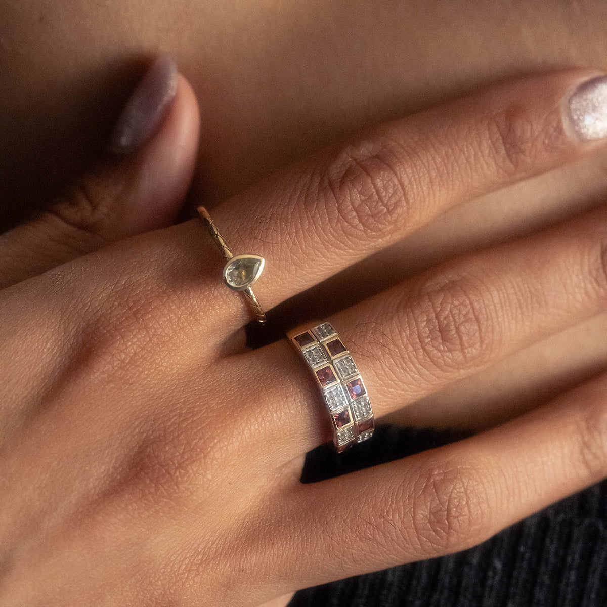 A manicured hand wears the Family Gold Solid Gold Garnet and Topaz Checkerboard Band, featuring a teardrop-shaped stone and a checkerboard band set with garnet and white topaz.