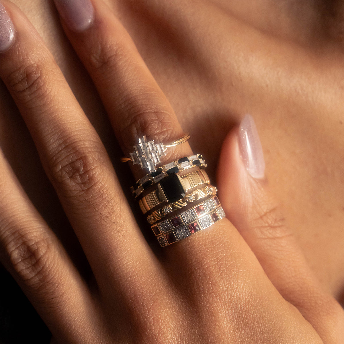 A close-up of manicured light pink nails wearing stacked gold and silver rings, including Family Gold’s Solid Gold Garnet and Topaz Checkerboard Band with geometric designs and colorful gemstones.