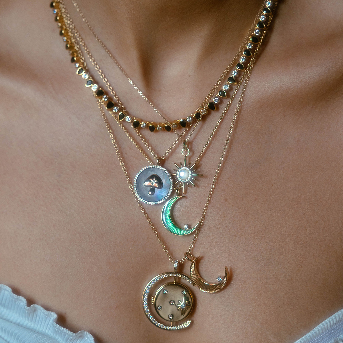 A close-up of a womans neck showcases the Luna Locket Necklace, a 14k gold plated piece with layered crescent moon, starburst, pearls, and beaded accents, styled over a white ribbed top.