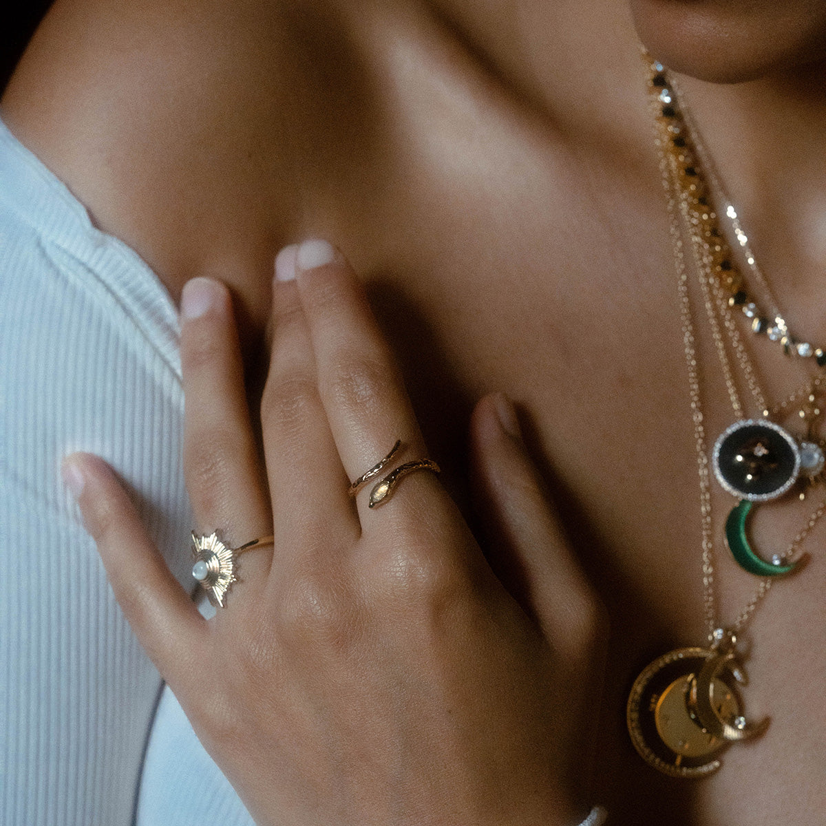Wearing layered gold necklaces with crescent moon and circular pendants and the Mystic Serpent Ring in Labradorite, plus other gold rings including a sun-shaped ring, this person rests their hand on their shoulder in a white ribbed off-shoulder top.