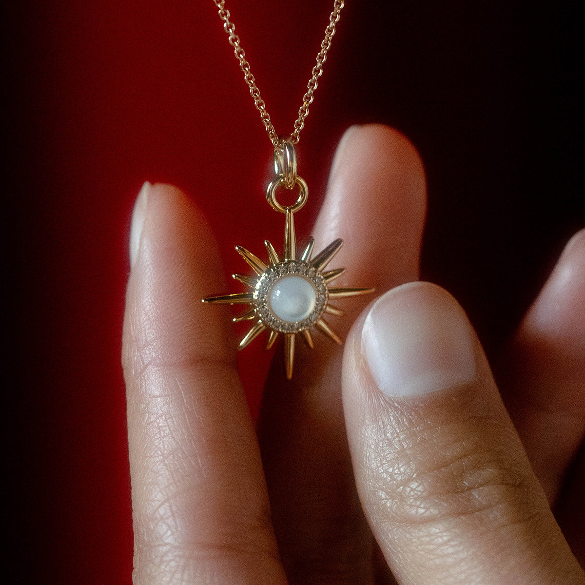 A hand holds the Charms by Elizabeth Stone sunburst pendant, featuring a round mother of pearl center on a delicate 14k gold plated chain, set against a dark red background.
