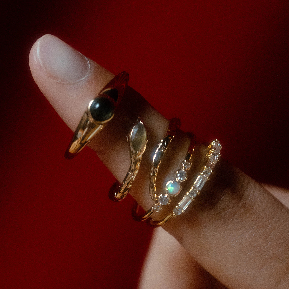 A close-up of a hand against a red background with five gold rings, including the Opal Stacking Ring, stacked on the tip of an upward-pointed finger.