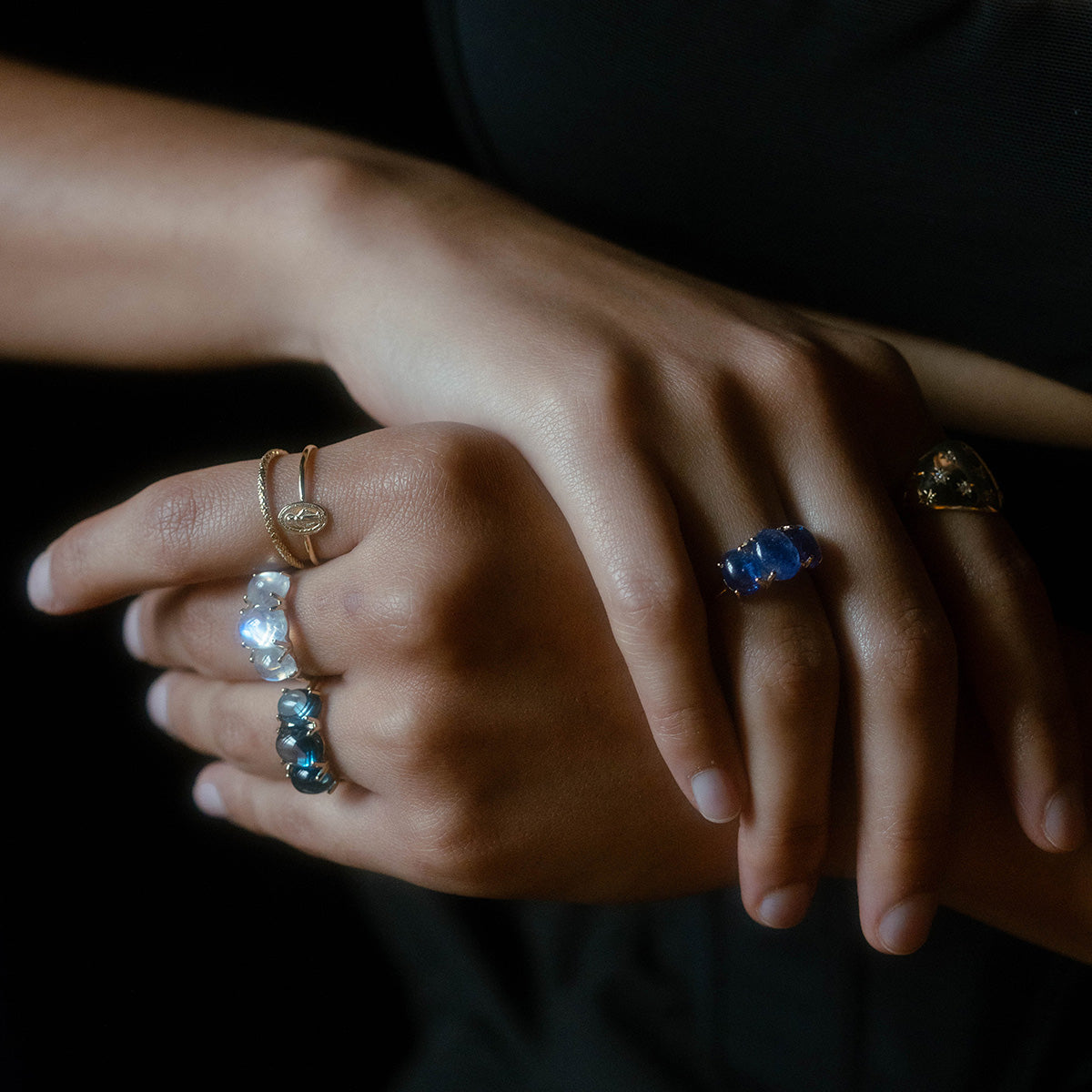 A close-up of two crossed hands wearing yellow gold rings with natural gemstones and bands, set against a dark background. The rings include blue, clear, and gold stones, featuring the striking Sweetest Gumdrop Ring.