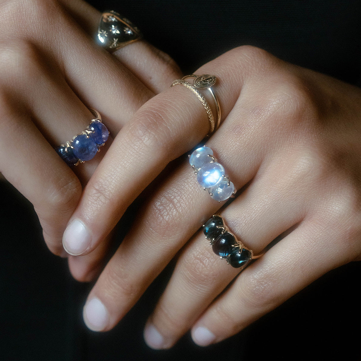 A close-up of two hands wearing yellow gold rings, including the Sweetest Gumdrop Ring set with natural blue, green, and clear gemstones, shown against a dark background.