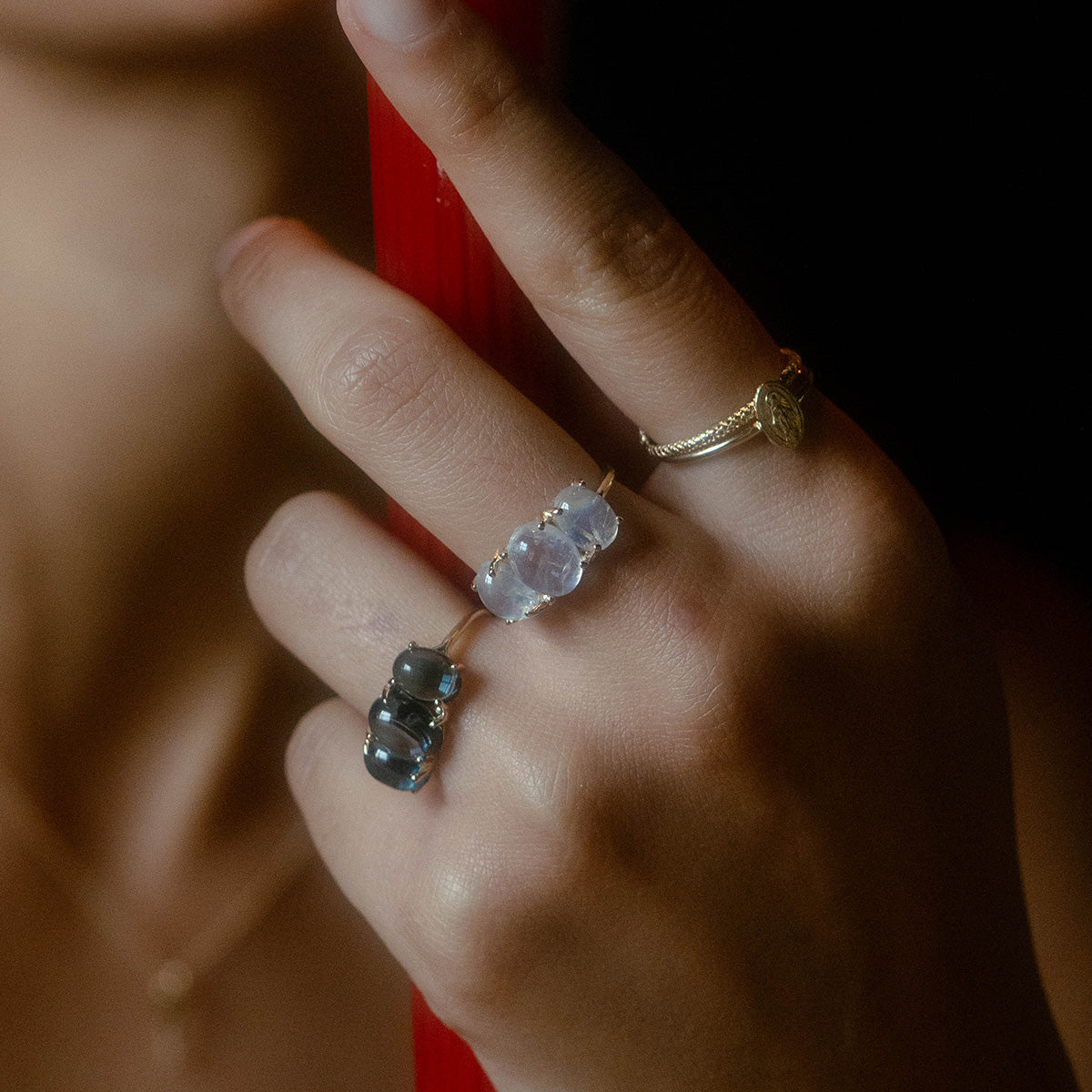 A close-up of a hand holding a red object, wearing three rings: the Sweetest Gumdrop Ring with translucent stones, one with dark gemstones, and a yellow gold ring featuring an engraved oval charm. The softly blurred background enhances the focus.