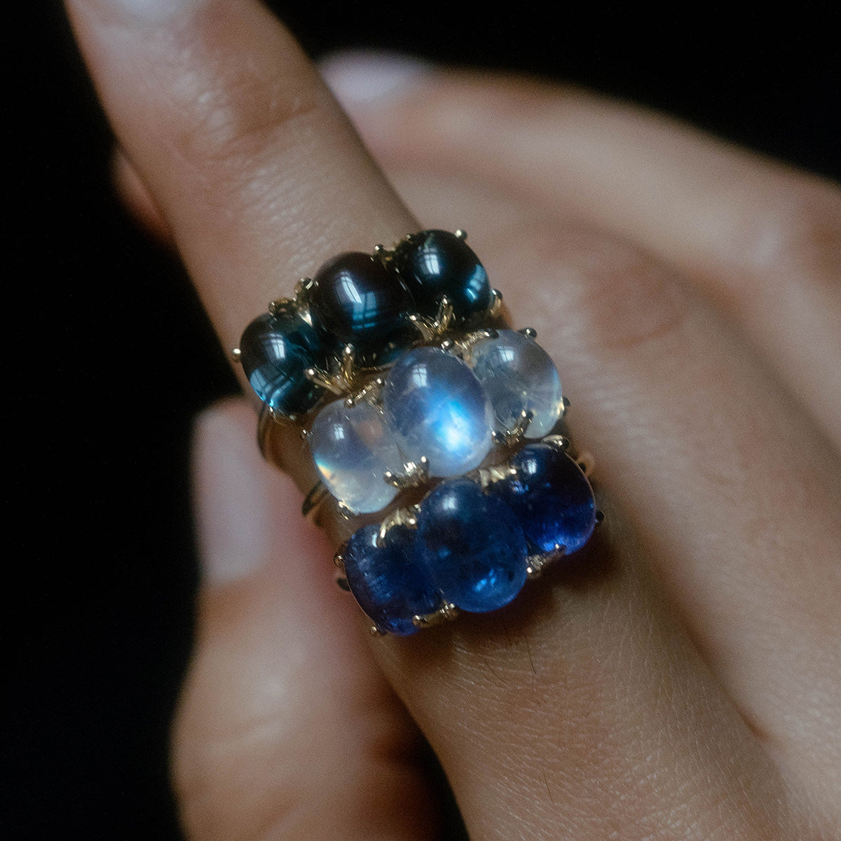 A hand wears three Sweetest Gumdrop Rings in yellow gold, stacked on one finger with natural cabochon gemstones—dark green, translucent white, and deep blue—set against a dark background.